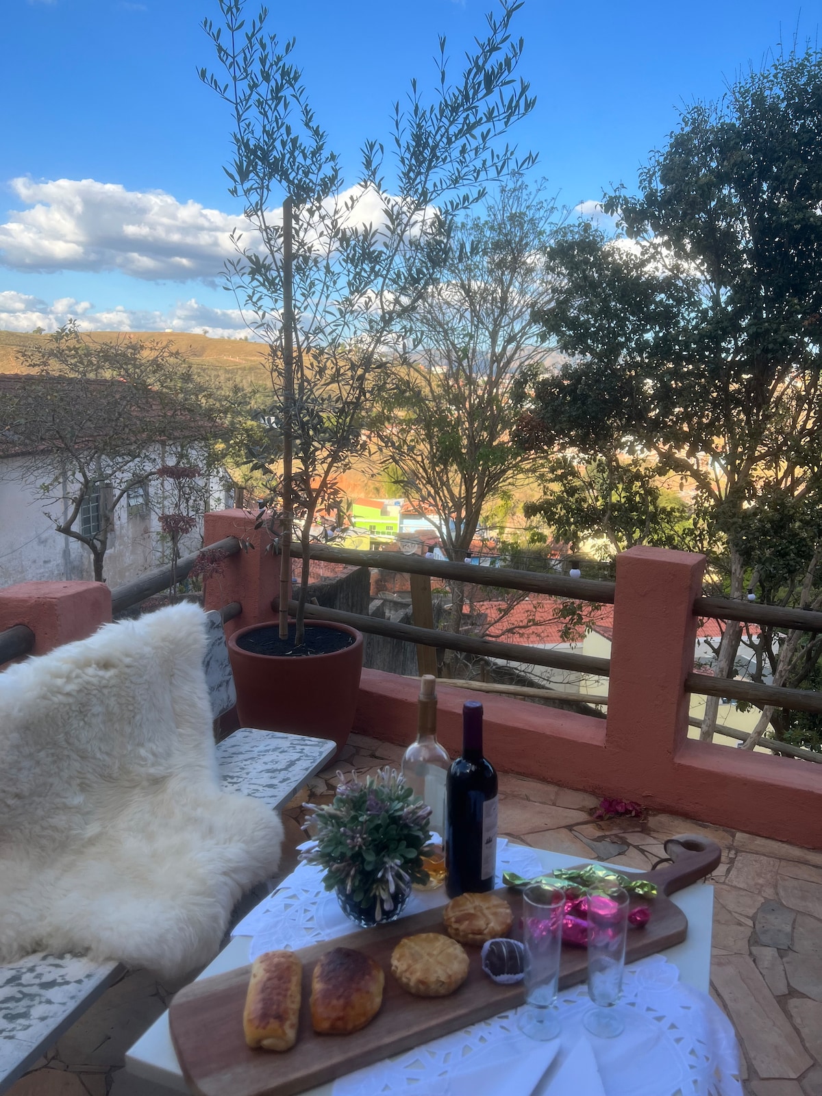 An outdoor space is depicted with a table set for dining, featuring a selection of baked goods, wine, and decorative glasses. Potted trees and a clear blue sky complete the scene. A railing provides a view of the surrounding area, enhancing the inviting ambiance.