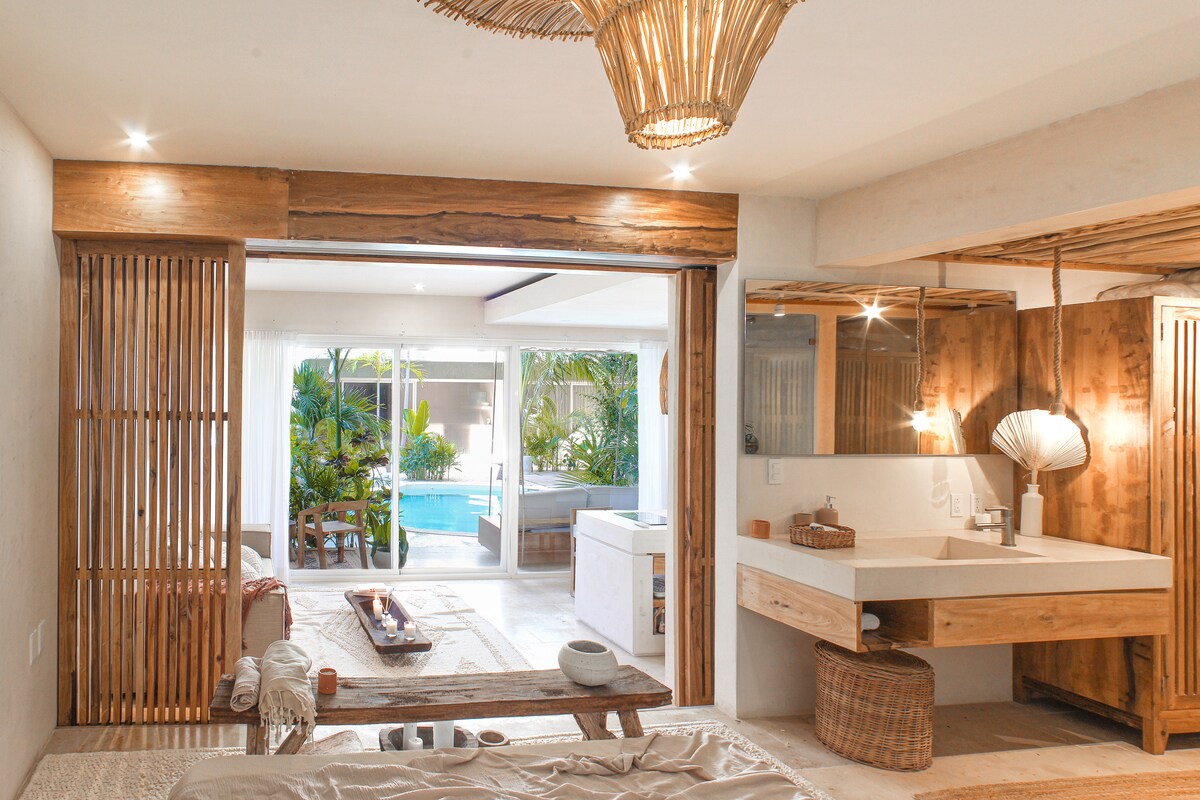 A spacious bedroom is illuminated by natural light from glass doors leading to a private patio. Wooden elements create a warm ambiance, while a minimalist bathroom area features a modern sink and round mirror. The view offers a glimpse of the inviting pool and lush greenery.