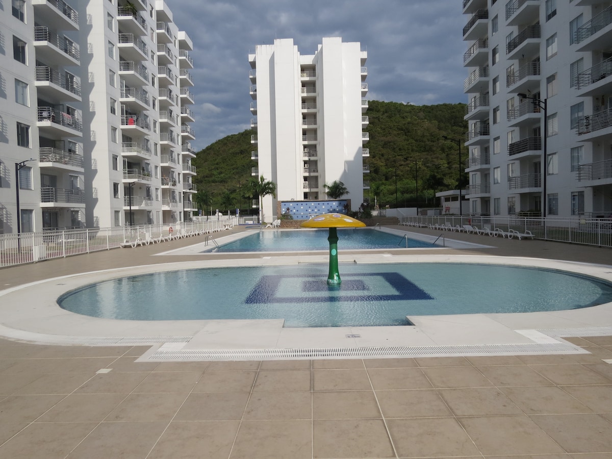 Two large swimming pools are visible, one oval and one rectangular, surrounded by tiled walkways. A colorful water feature stands at the center of the circular pool. Apartment buildings rise in the background against a hilly landscape under a partly cloudy sky.