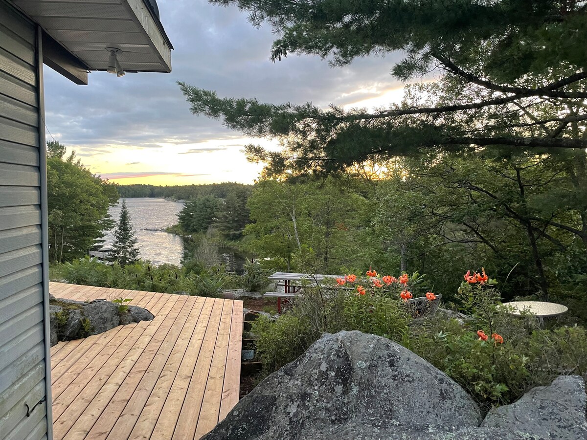 A wooden deck extends from the cottage, bordered by natural rock formations and vibrant orange flowers. In the distance, a serene body of water reflects the soft colors of the sunset, framed by lush green trees and clouds in a changing sky.