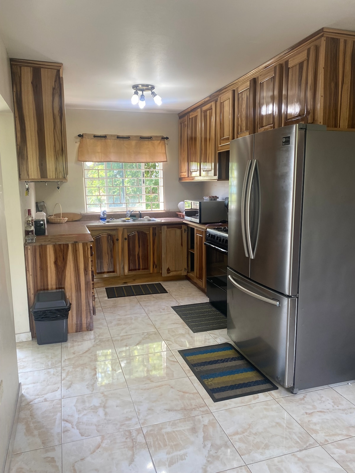 The kitchen showcases wooden cabinetry with a smooth countertop, complemented by modern stainless-steel appliances. Natural light enters through a window adorned with a light fabric shade, while decorative mats enhance the tiled floor. A waste bin is discreetly positioned near the entrance.