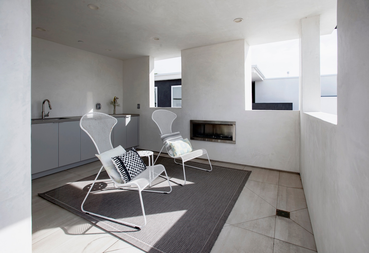 A covered outdoor lounge area features two white chairs positioned on a light-colored rug. A sleek modern fireplace is built into the wall, with a minimalist kitchen area visible in the background. Natural light filters through the open windows, creating a bright and airy space.