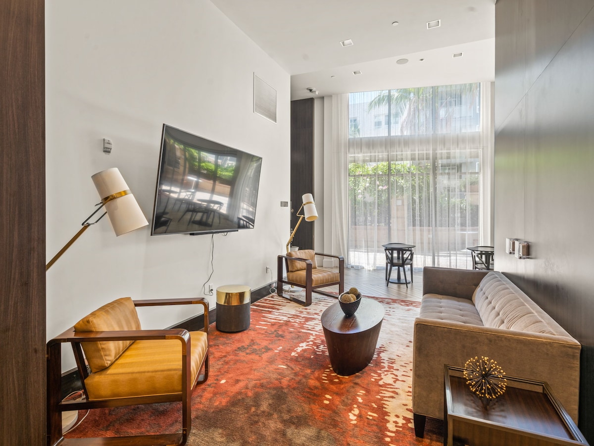 A comfortable lounge area features a large flat-screen TV mounted on the wall. Two stylish armchairs and a plush sofa are arranged around a low coffee table. Natural light filters through sheer curtains, illuminating the warm-toned rug beneath.