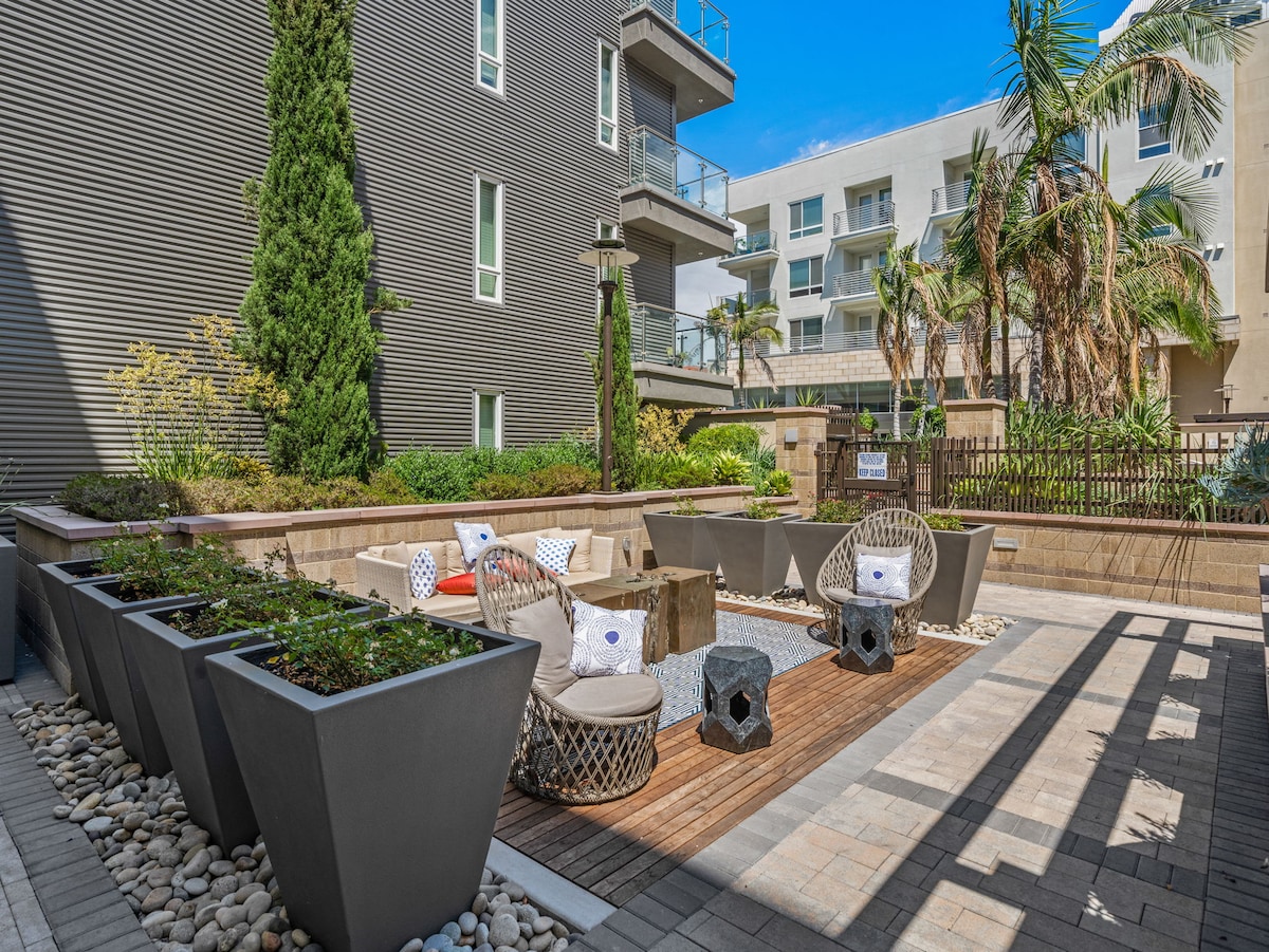 An outdoor lounge area is set with comfortable seating arranged around a wooden deck. Decorative planters house greenery, while pathways are lined with smooth stones. Tall palms provide a tropical feel, and modern architecture serves as a backdrop to this inviting space.