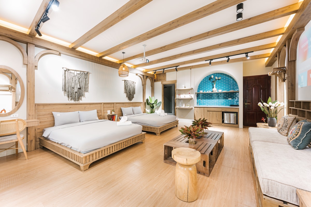 The spacious area features two queen beds with natural wood frames, complemented by light-colored bedding. A comfortable seating arrangement is positioned near the wall, and plants add a touch of greenery. A modern kitchenette with a blue-tiled backsplash is visible, along with wood accents throughout.