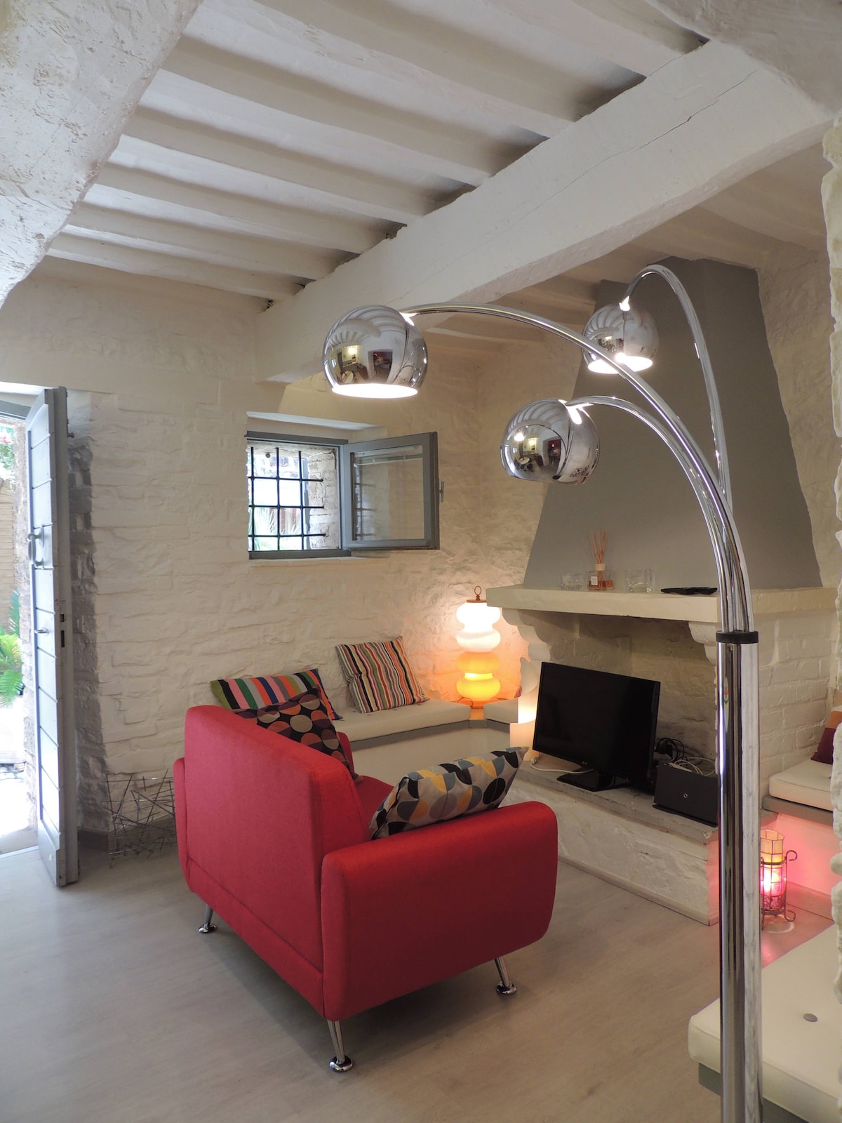 A vibrant living area features a red sofa with decorative cushions and a sleek floor lamp. A small television is positioned next to a whitewashed fireplace. Natural light enters through a window and a nearby door, which leads to an outdoor space.