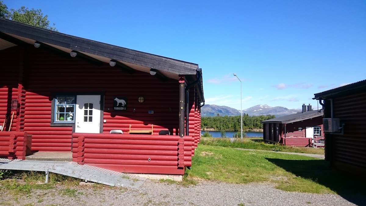 A red log cabin with a wooden porch is shown against a clear blue sky. Surrounding green grass and gravel paths lead to the cabin's entrance. The view includes distant mountains, and a body of water is visible nearby, enhancing the natural setting.