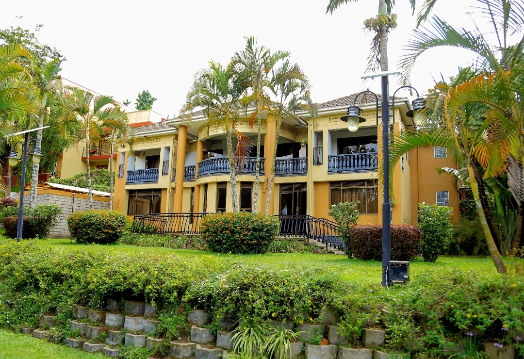 The hotel exterior features a two-story building surrounded by lush greenery and palm trees. Balconies with blue railings are visible on the upper level, while well-maintained garden areas frame the property, providing a welcoming environment for guests.