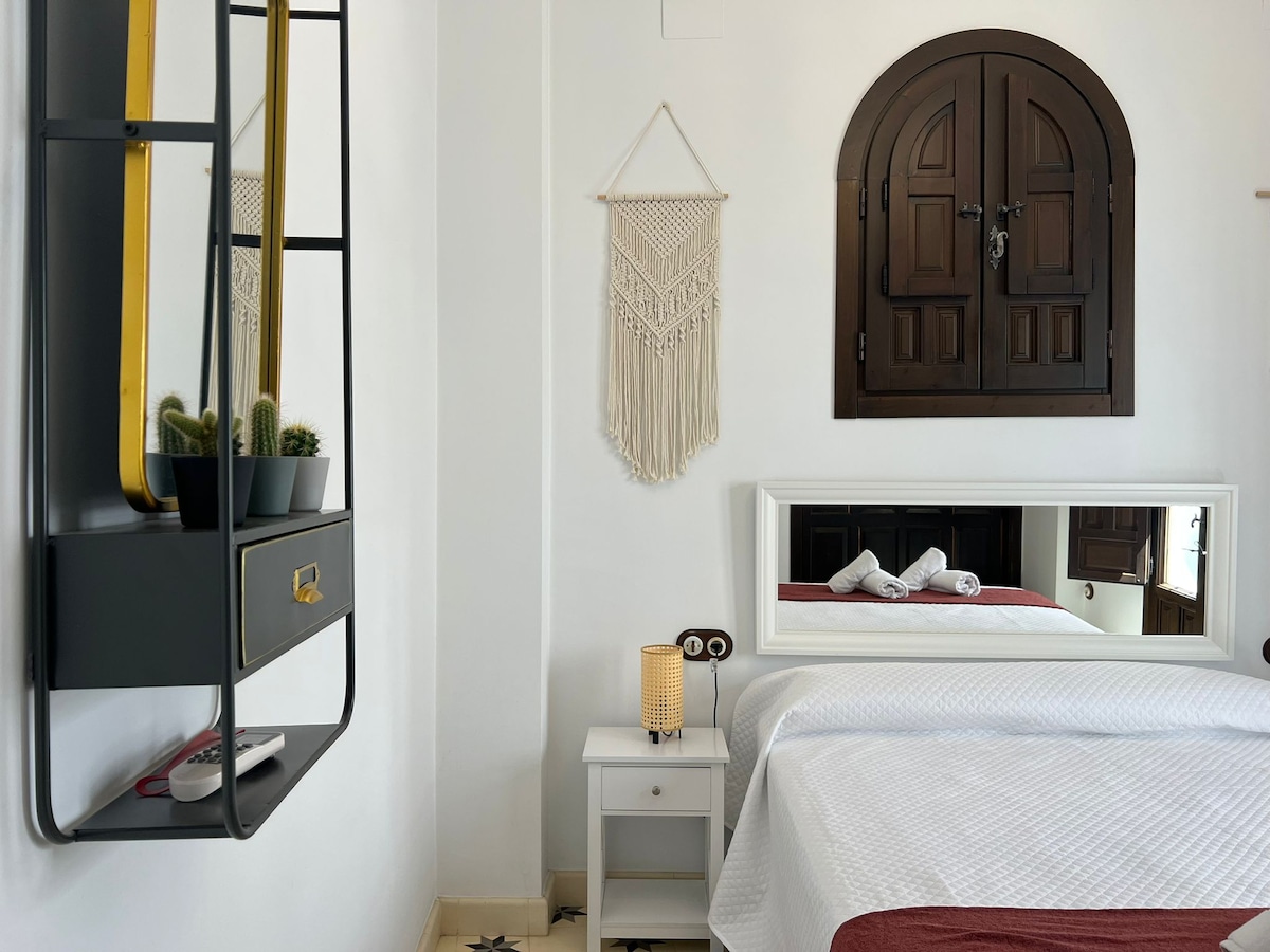 A well-appointed bedroom features a spacious bed with fresh linens. A large mirror reflects the light from a nearby decorative lamp. A wall-mounted shelf holds small green plants, while a macramé wall hanging adds texture. Dark wood accents frame the archway of a doorway.