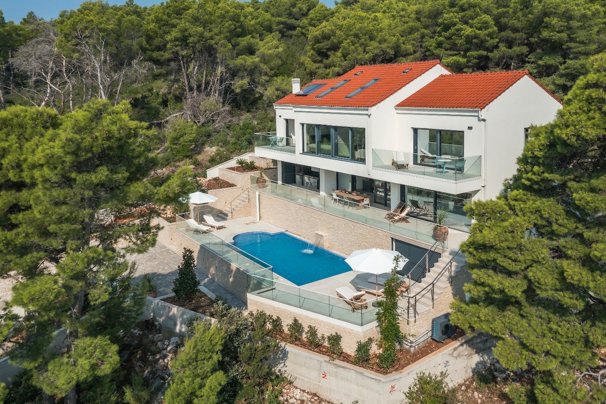 A contemporary villa stands amidst lush greenery, featuring a private saltwater pool surrounded by a spacious terrace. The exterior showcases large glass windows and multiple seating areas, offering inviting spaces for relaxation and outdoor dining.