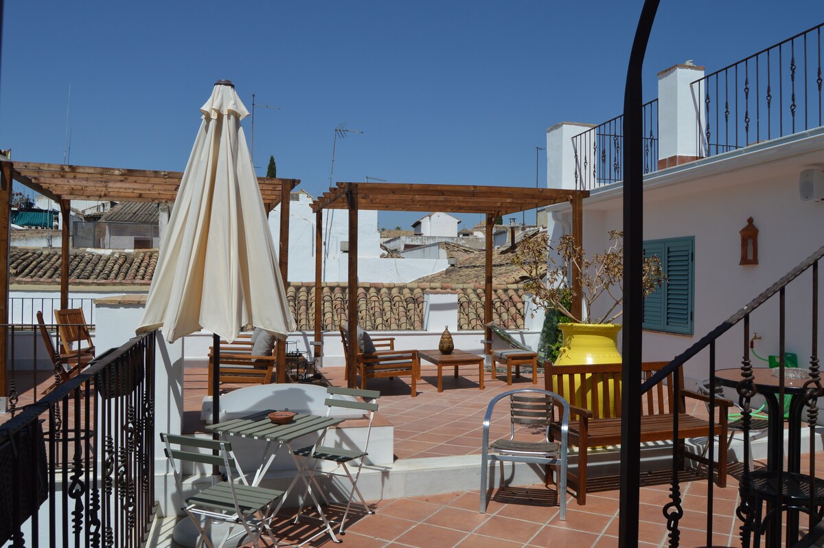 A spacious terrace is featured, showcasing wooden pergolas providing shade. Lounge chairs and a dining area with metal chairs and a table are arranged for relaxation. A vibrant yellow planter adds a pop of color to the space, with traditional tile and clear blue skies visible in the background.