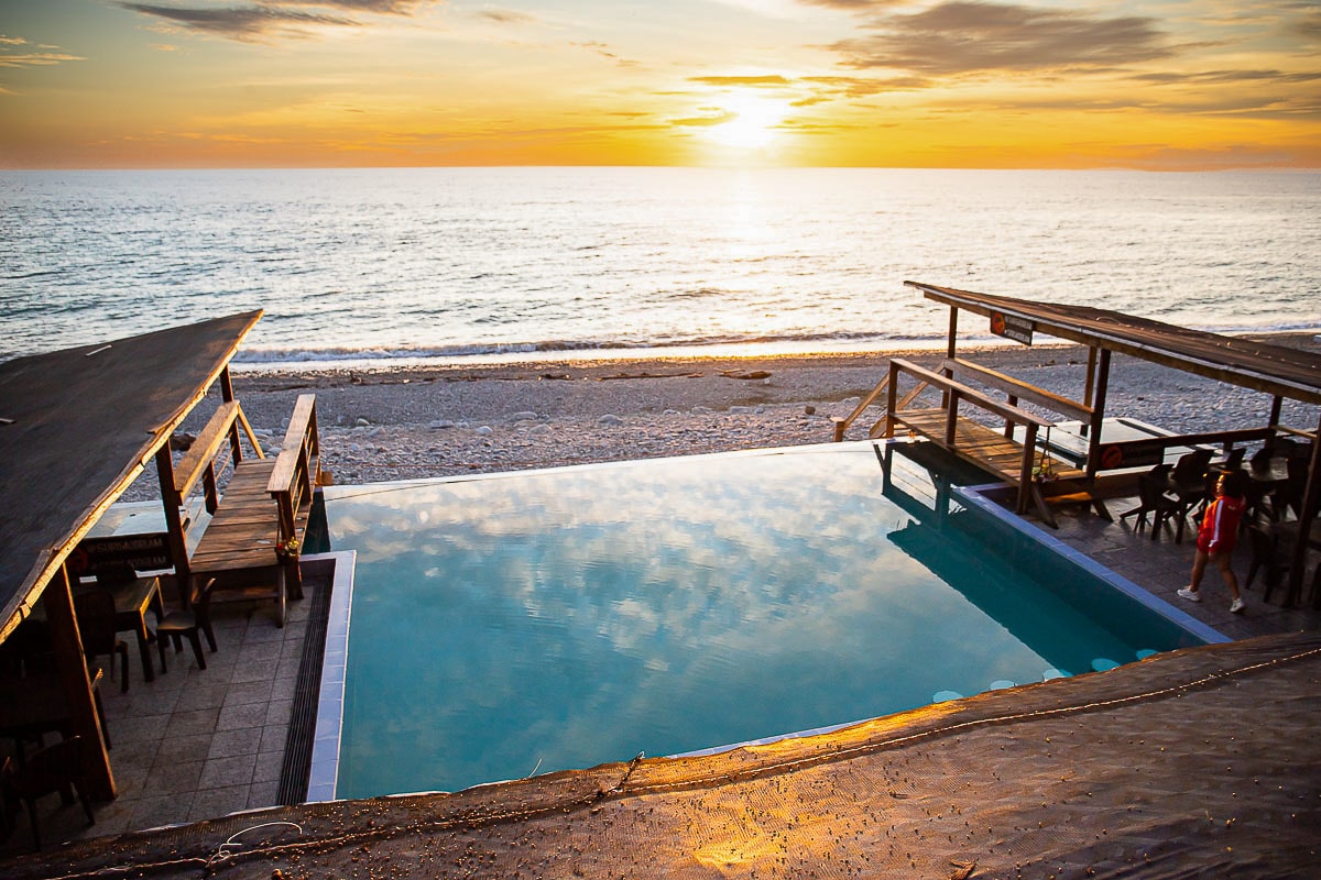 A tranquil swimming pool reflects the sunset over the ocean, with smooth wooden decking surrounding it. Beach chairs and tables are positioned under shaded structures, offering a serene view of the pebble beach and calm sea.