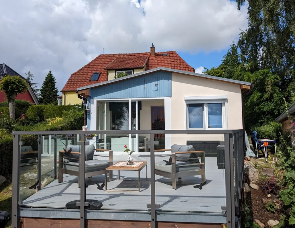 The bungalow exterior is presented, showcasing a spacious terrace with seating arrangements and a central table. Large glass doors provide access to the interior, while the house features a mix of muted and vibrant colors under a partly cloudy sky.