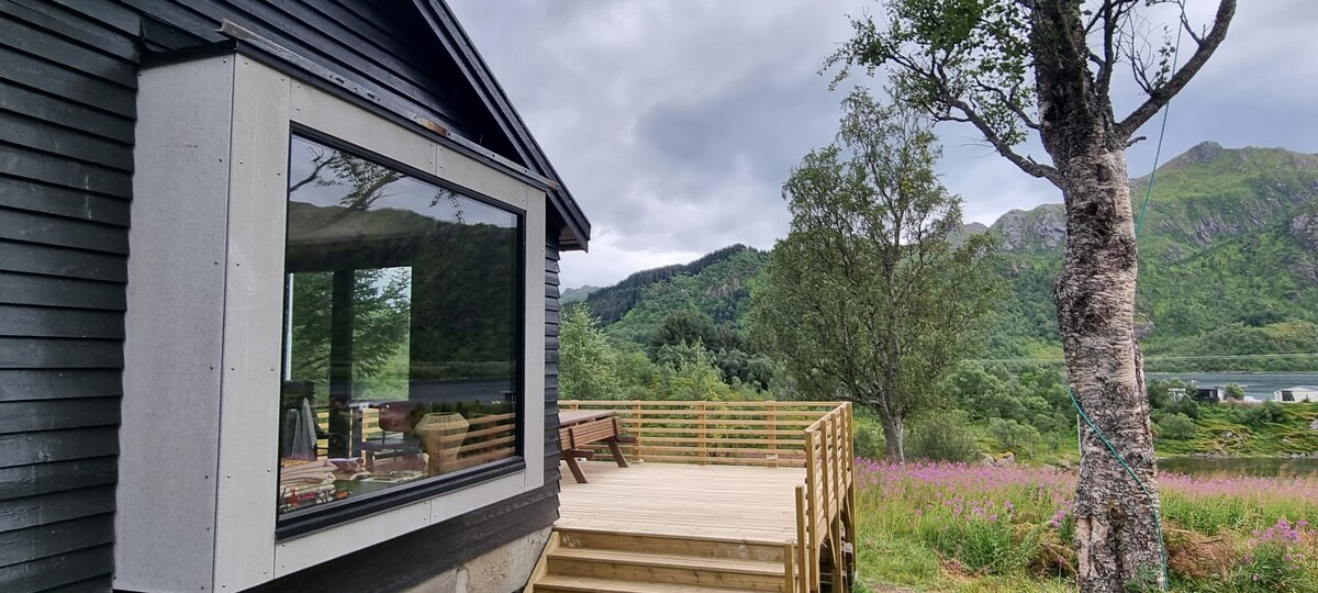 A spacious terrace offers wooden decking adjacent to a modern building, with large glass windows allowing for views of the surrounding green hills and natural landscape. A tree stands nearby, contributing to the serene environment.