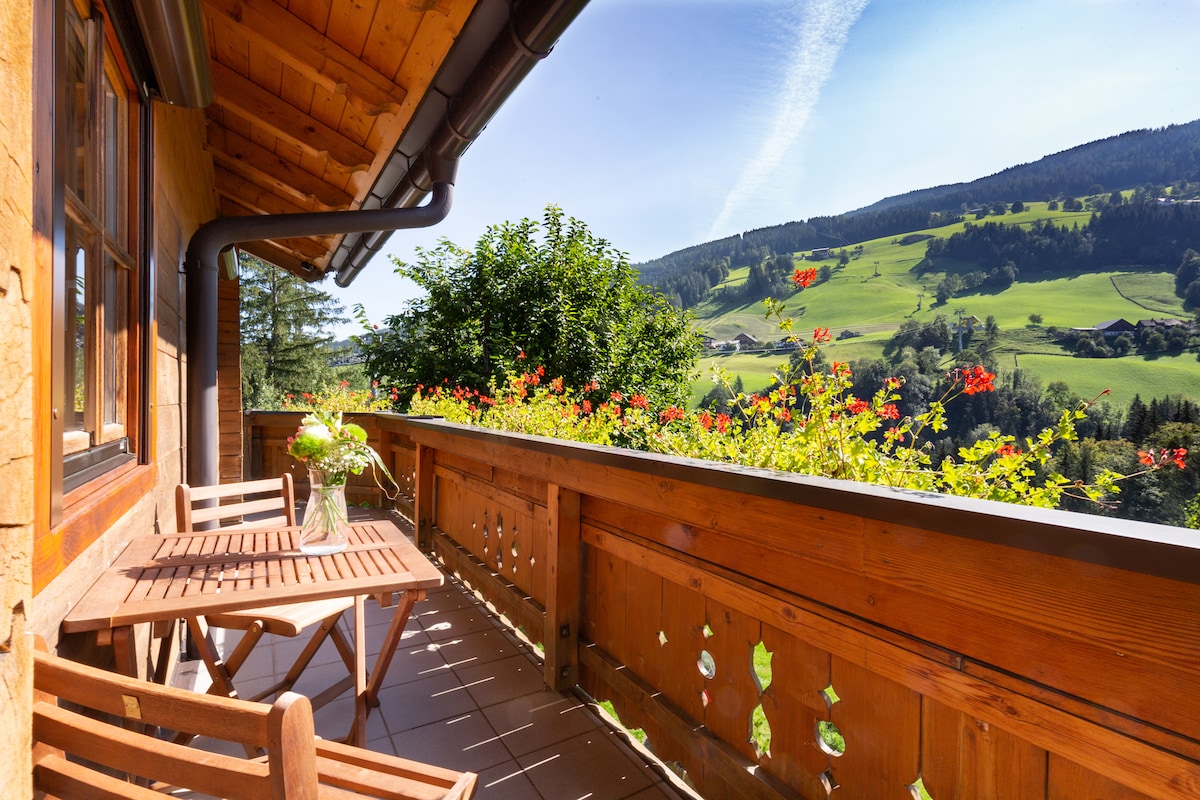 A wooden balcony features a table with chairs, overlooking a scenic landscape of green hills and blooming flowers. The sunlight illuminates the space, enhancing the natural beauty of the surrounding area.