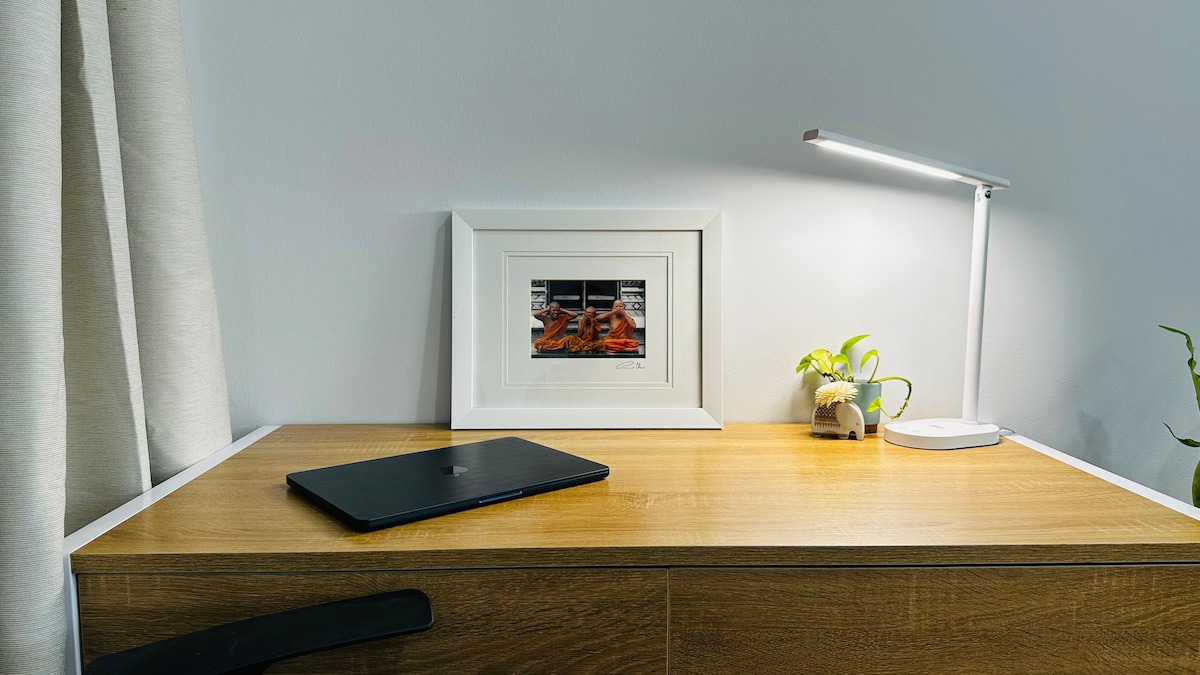 A neatly arranged workspace features a sleek laptop on a wooden surface. A framed art piece with a warm color palette provides visual interest, while a small potted plant and a modern desk lamp enhance the ambiance.