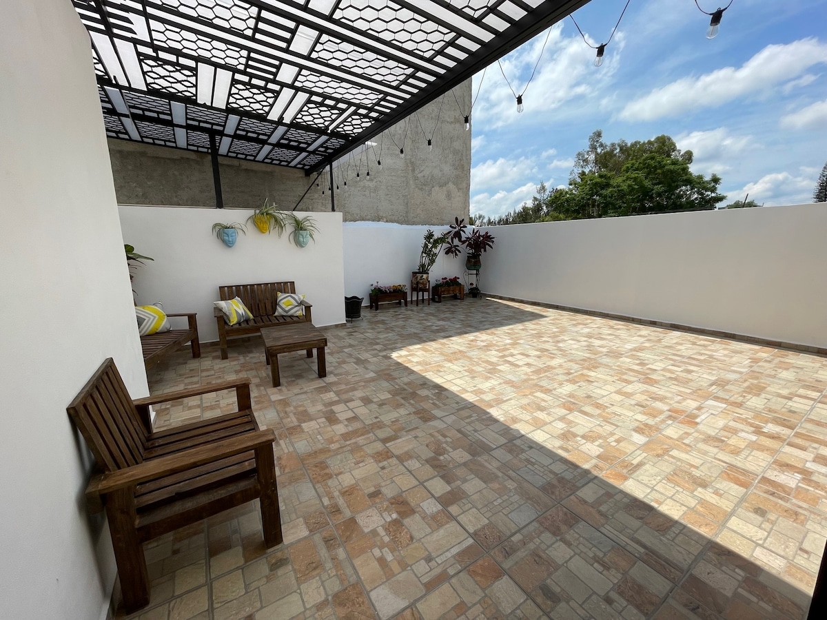 A spacious terrace is presented, featuring a tiled floor and a covered roof adorned with decorative lights. Wooden seating arrangements are positioned on one side, accompanied by colorful cushions. Lush greenery is visible in the background, providing a relaxing environment.
