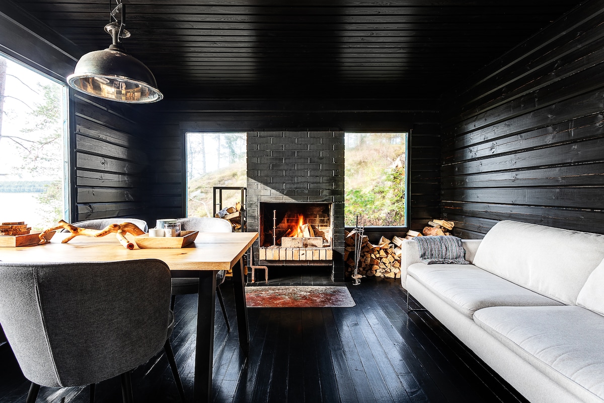 A cozy living space is presented, featuring a wood-burning fireplace surrounded by neatly stacked firewood. A modern dining table is positioned adjacent to a light grey sofa, with large windows allowing natural light to fill the room and offer views of the surrounding landscape.