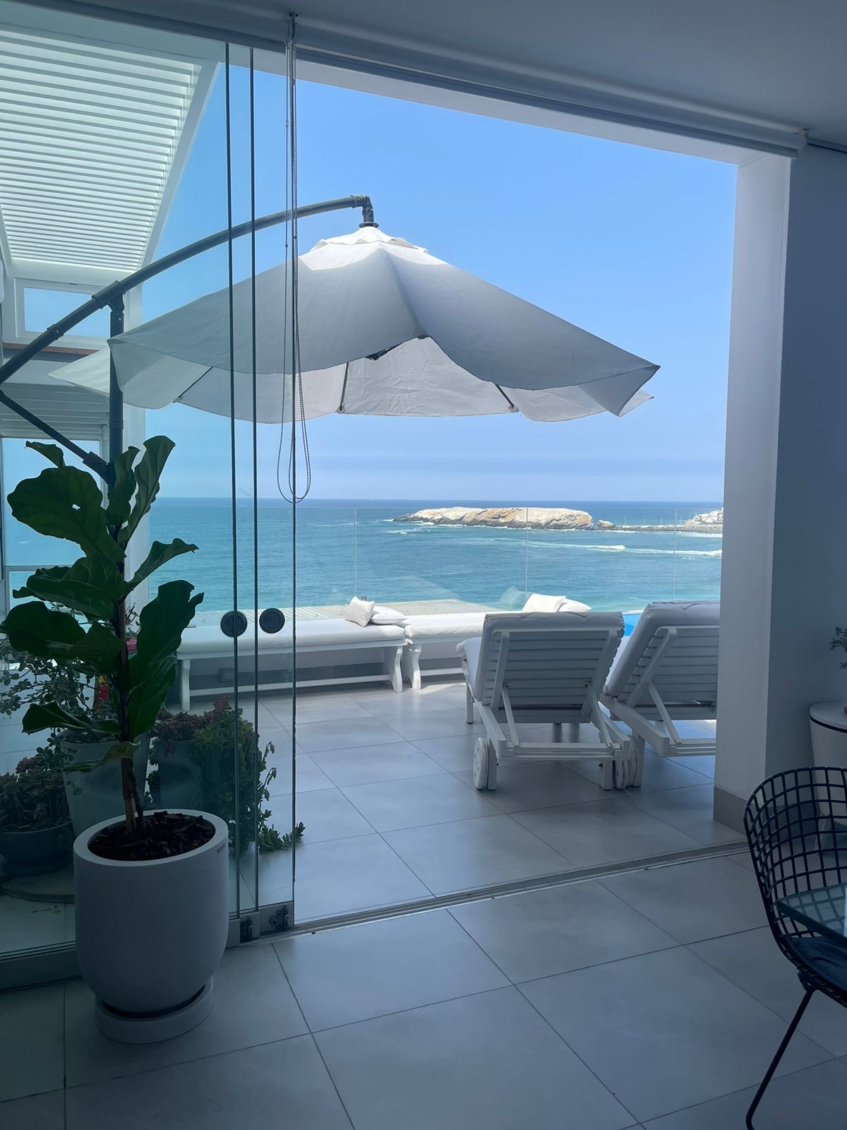 A sunny outdoor terrace features lounge chairs overlooking the ocean, with an umbrella providing shade. Lush greenery is seen in a pot, adding a touch of nature. The sliding glass doors create a seamless transition between indoor and outdoor spaces.