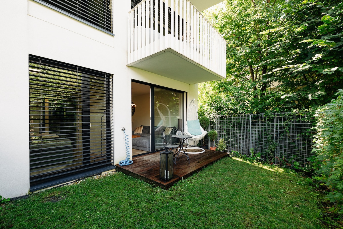 A patio area is visible, featuring a small wooden deck with a round table and two chairs. A stylish hanging chair can be seen, surrounded by greenery. The exterior walls of the building are light-colored, with large windows framed by dark accents, providing a serene atmosphere.