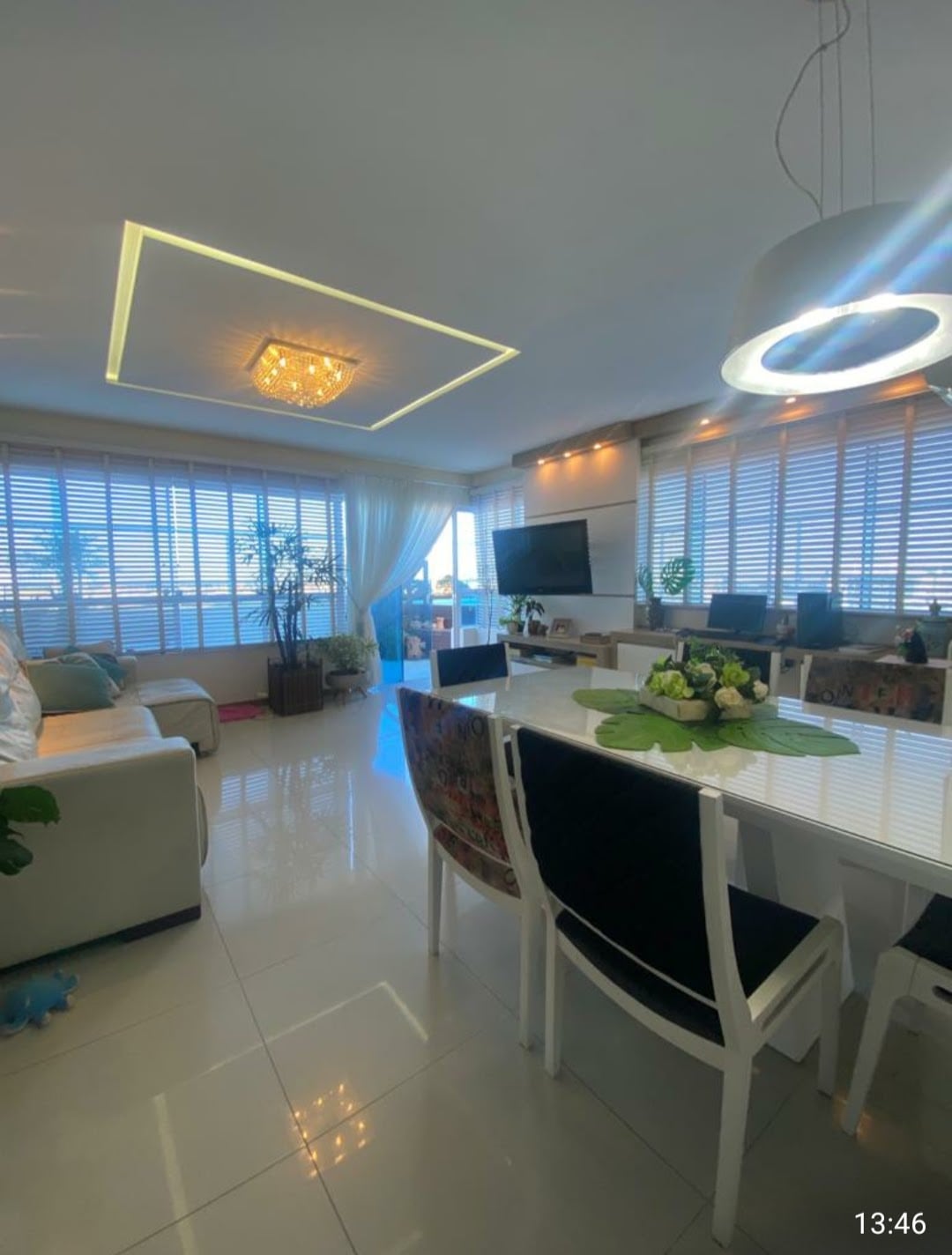 A bright living space showcases a modern design with floor-to-ceiling windows allowing natural light to fill the area. A comfortable seating arrangement is complemented by a large television and decorative plants. A dining table with elegant chairs sits nearby, enhancing the open layout.