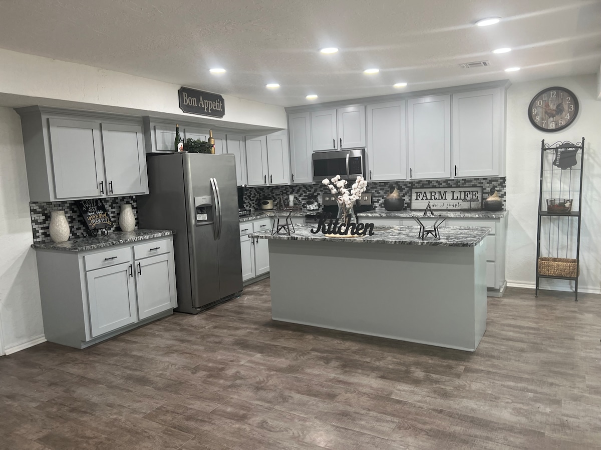 A modern kitchen features light grey cabinetry and a central island with bar seating. Stainless steel appliances and a decorative backsplash enhance the functionality. A clock adorns the wall above a shelving unit, while a 'Kitchen' sign adds a welcoming touch.