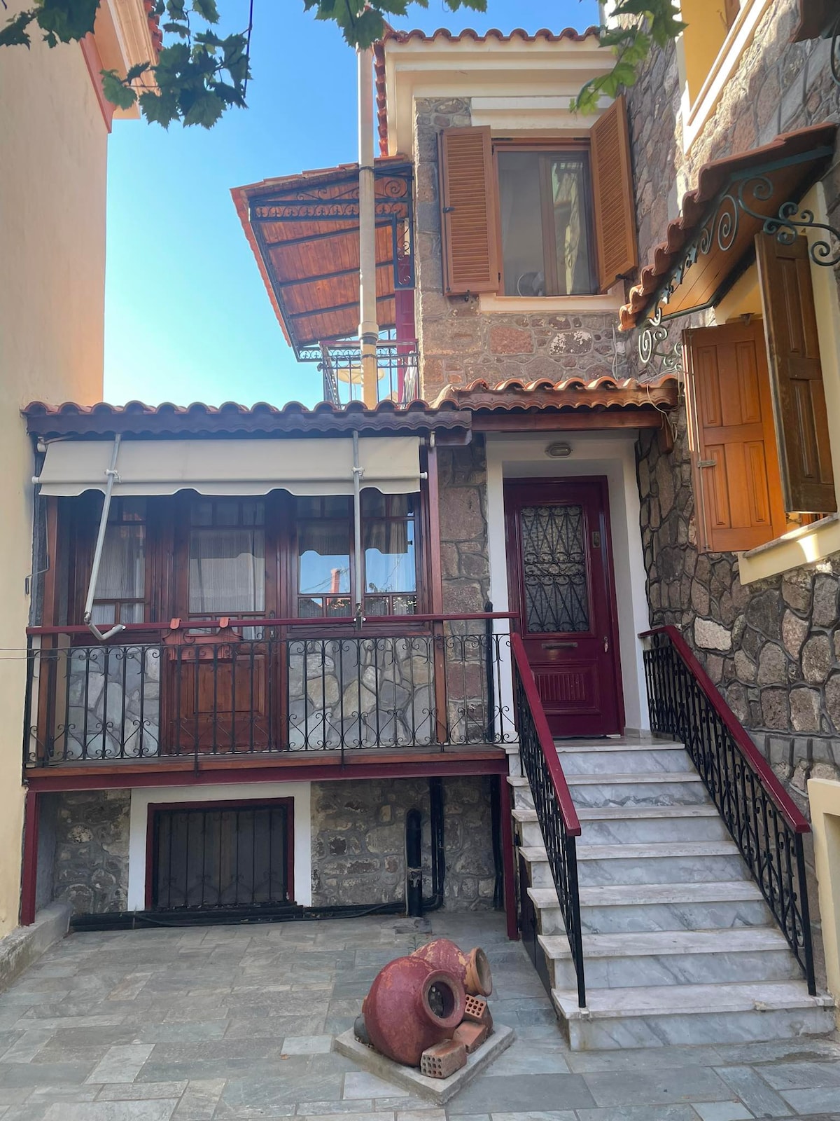 A charming stone facade showcases the entrance to Melianto House, featuring a set of stone steps leading up to a dark wooden door. A small balcony with wooden shutters and a wrought iron railing enhances the welcoming exterior, complemented by a traditional clay pot resting on the ground.