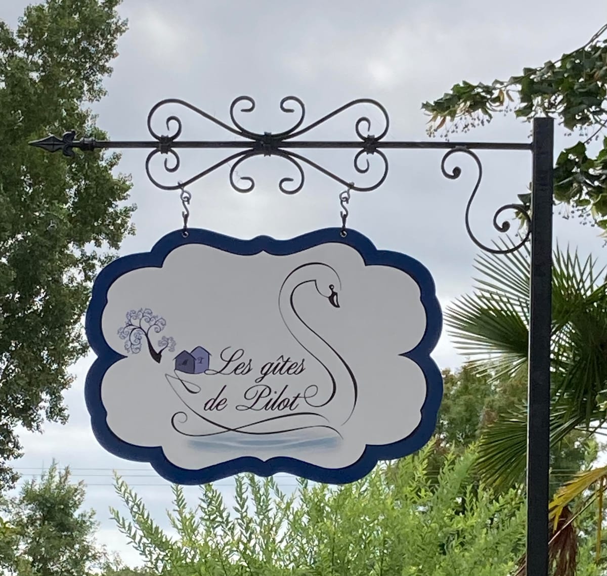 A decorative sign hangs from a wrought iron frame, featuring the name 'Les gîtes de Pilot.' The design includes an elegant swan and a small house, surrounded by lush greenery and palm leaves in the background.