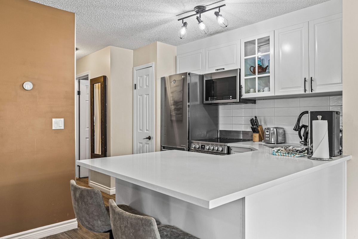 A modern kitchen space is highlighted by sleek cabinetry and stainless steel appliances. A large island with seating for two offers an inviting area for casual dining or conversation. Natural light fills the room, showcasing the contemporary finishes and open layout.
