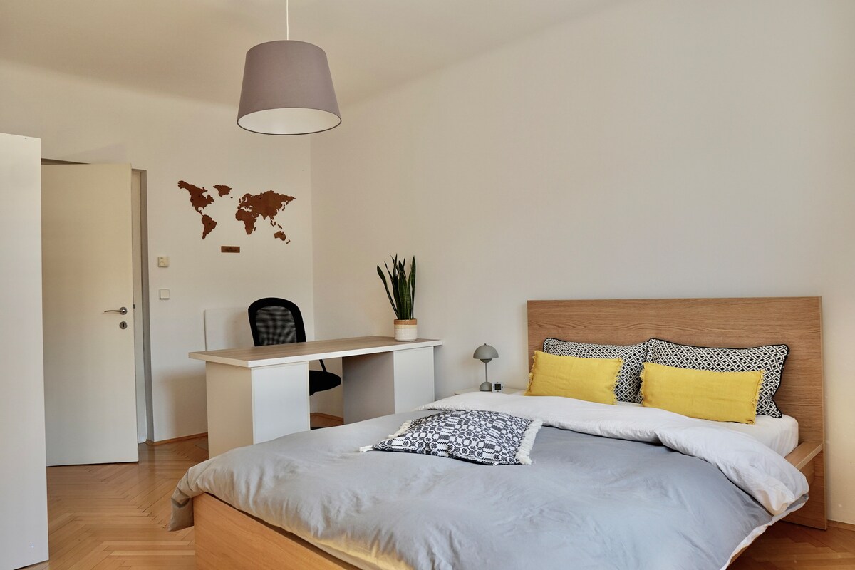 A light-filled bedroom features a spacious bed with gray bedding and two yellow pillows. A desk is positioned nearby, complemented by a black office chair. A decorative world map is mounted on the wall, and a potted plant adds a touch of greenery.