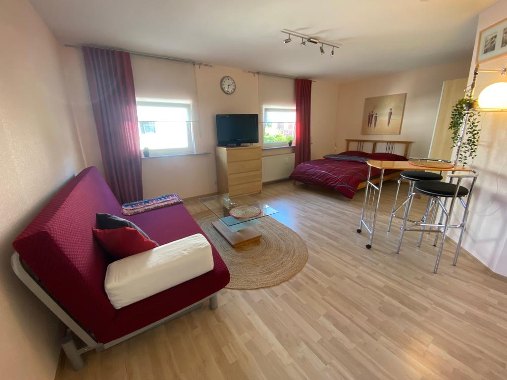 An open living space features a red seating area with white cushions, a wooden dining set, and a television on a dresser. Two beds are arranged with red bedding, while a circular rug is positioned centrally on the light-colored floor. Natural light streams through the windows.