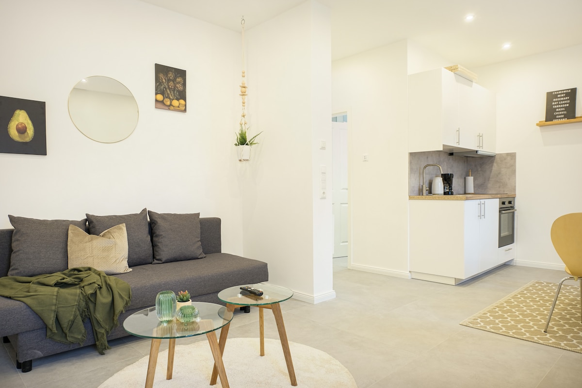 A modern living area is presented with a gray sofa adorned with decorative cushions. Two circular coffee tables sit atop a soft area rug. Natural light fills the space, highlighting the minimalist kitchen with appliances and accents of art on the walls.
