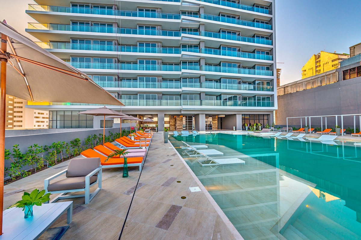 The oceanfront pool area features several lounge chairs arranged along the water's edge. A modern building stands in the background, with multiple floors visible. Umbrellas provide shade over some seating, while surrounding greenery adds a touch of nature.