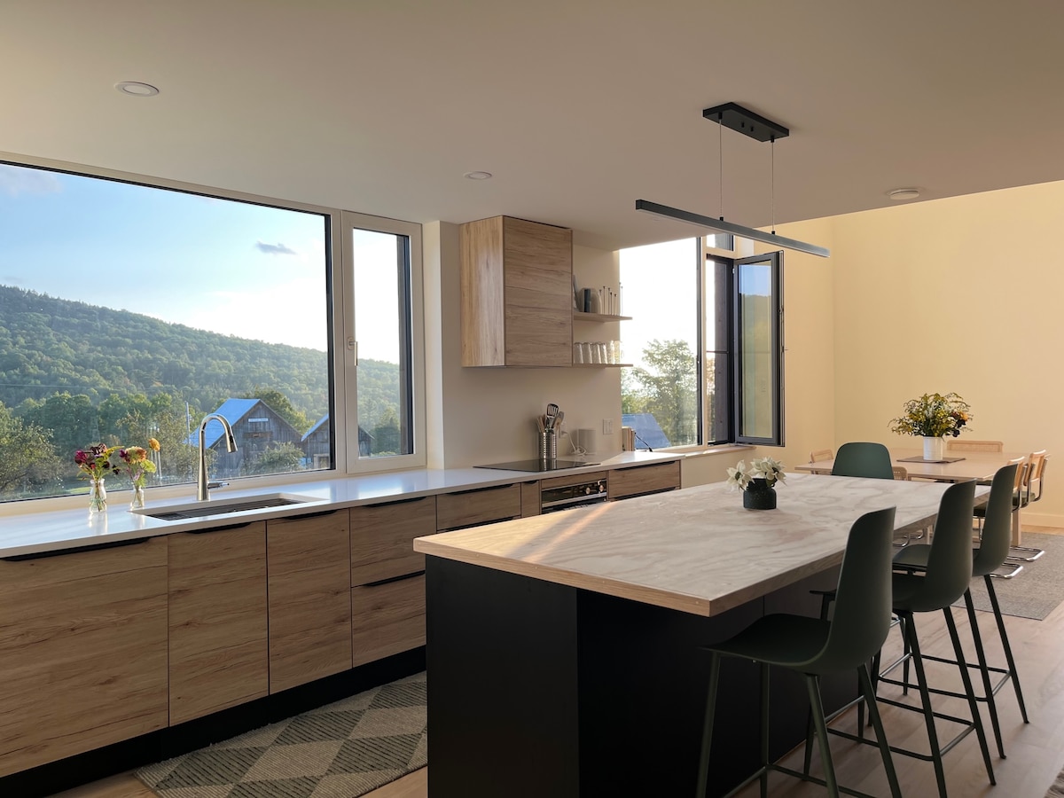 A bright kitchen features modern cabinetry and a spacious island with seating. Large windows reveal views of the surrounding hills, allowing natural light to fill the room. A simple floral arrangement adds a touch of freshness to the countertop.