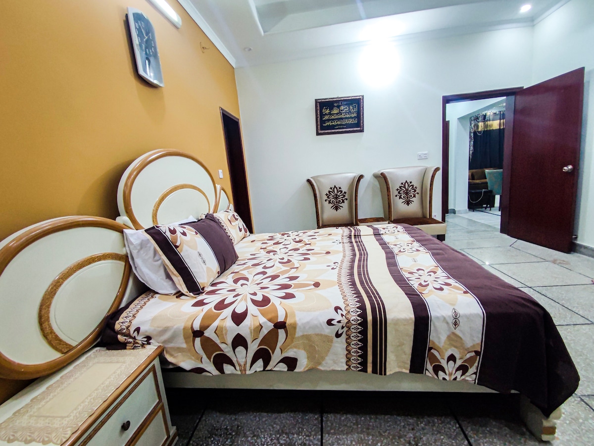 A bedroom features a spacious bed with a patterned brown and cream bedspread. Two upholstered chairs are positioned nearby, and a bedside table stands beside the bed. The walls are painted in warm tones, and a decorative wall piece is visible.