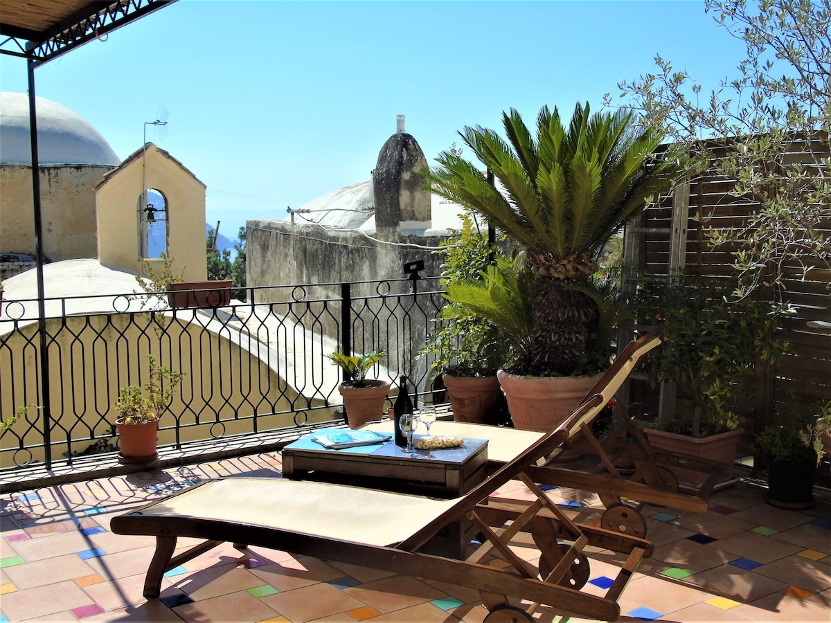 A spacious terrace is shown, featuring a wooden lounge chair and a small table. Potted plants add greenery to the area. A view of charming rooftops and the sky can be seen in the background, creating a relaxed outdoor space.