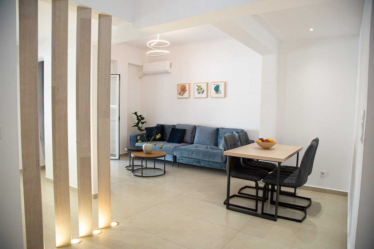 A modern living space is showcased, featuring a blue sofa arranged with decorative cushions. A dining table is paired with metal chairs, and a fruit bowl sits atop it. Wall art in muted tones adorns the walls, while natural light brightens the area through a nearby door.