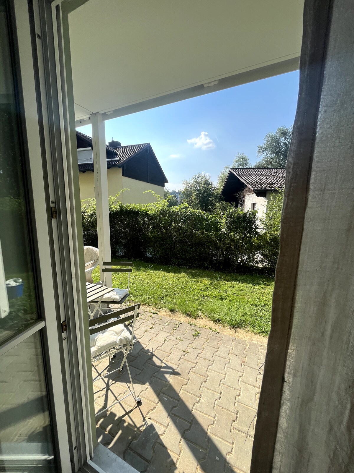 A view from the doorway reveals a private outdoor patio area with comfortable seating. Green grass and shrubs frame the space, while sunlight creates a bright and airy atmosphere. Nearby buildings can be seen in the background, adding to the serene setting.
