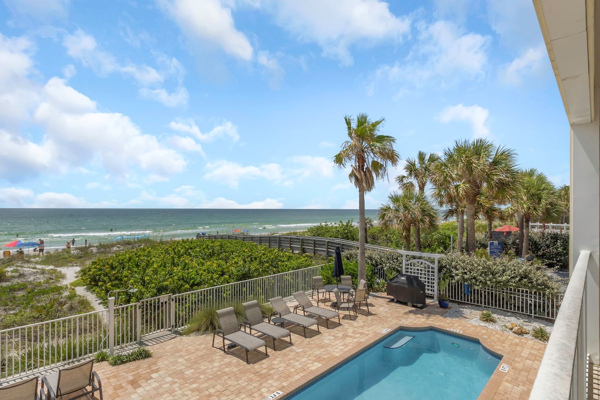 An inviting view of an outdoor area features a heated pool surrounded by lounge chairs. Lush greenery and palm trees frame the scene, leading to the sandy beach beyond. The ocean stretches out in the background under a clear blue sky, enhancing the coastal ambiance.