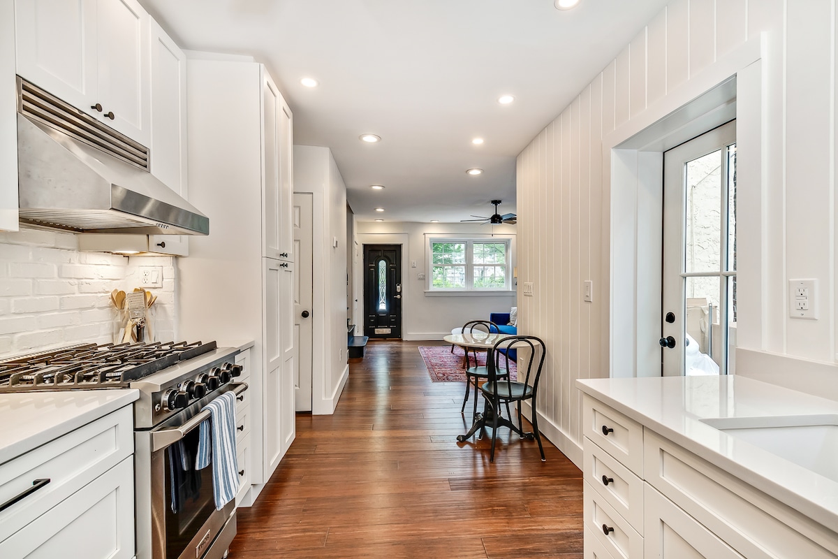 A modern kitchen presents crisp white cabinetry and stainless steel appliances, including a gas stove. Wood flooring extends into a well-lit dining area, featuring a round table with black chairs. Natural light enters through a sliding glass door, leading to an outdoor space.