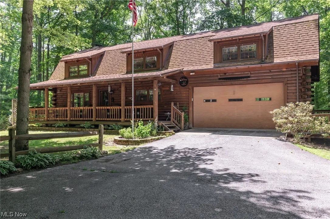 A spacious log cabin is nestled among lush trees, featuring a welcoming front porch with wooden railings. The natural-colored façade is complemented by large windows and a garage door, offering an inviting entrance to the secluded getaway.