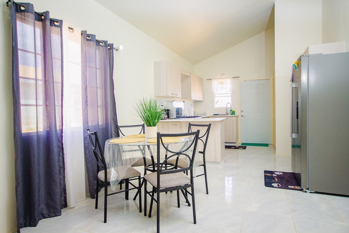 The spacious kitchen area features a round dining table surrounded by four chairs. Soft natural light enters through the sheer curtains, highlighting the modern cabinetry and appliances. A large refrigerator stands prominently against the wall, complementing the overall functionality of the space.
