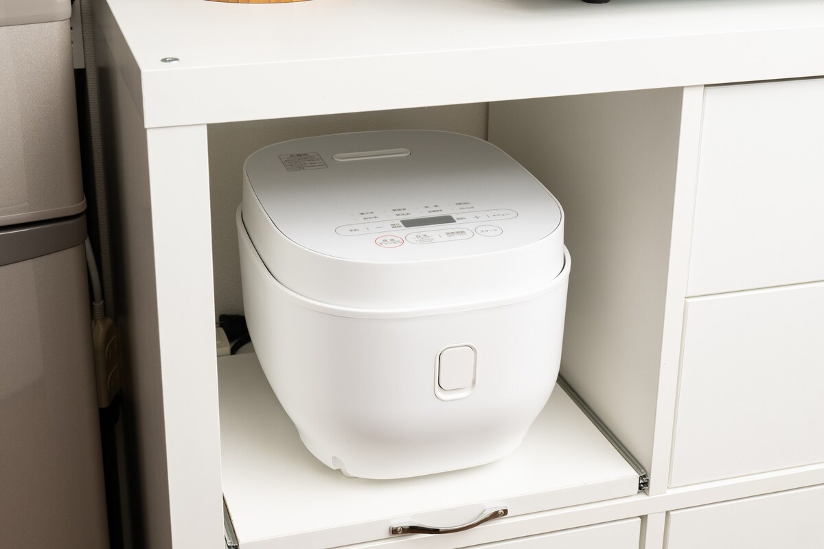 A rice cooker is neatly placed on a shelf within a white cabinet. The appliance features a clean design, with control buttons visible on its top surface, indicating its functionality. The surrounding space is organized, enhancing the kitchen's overall tidiness.