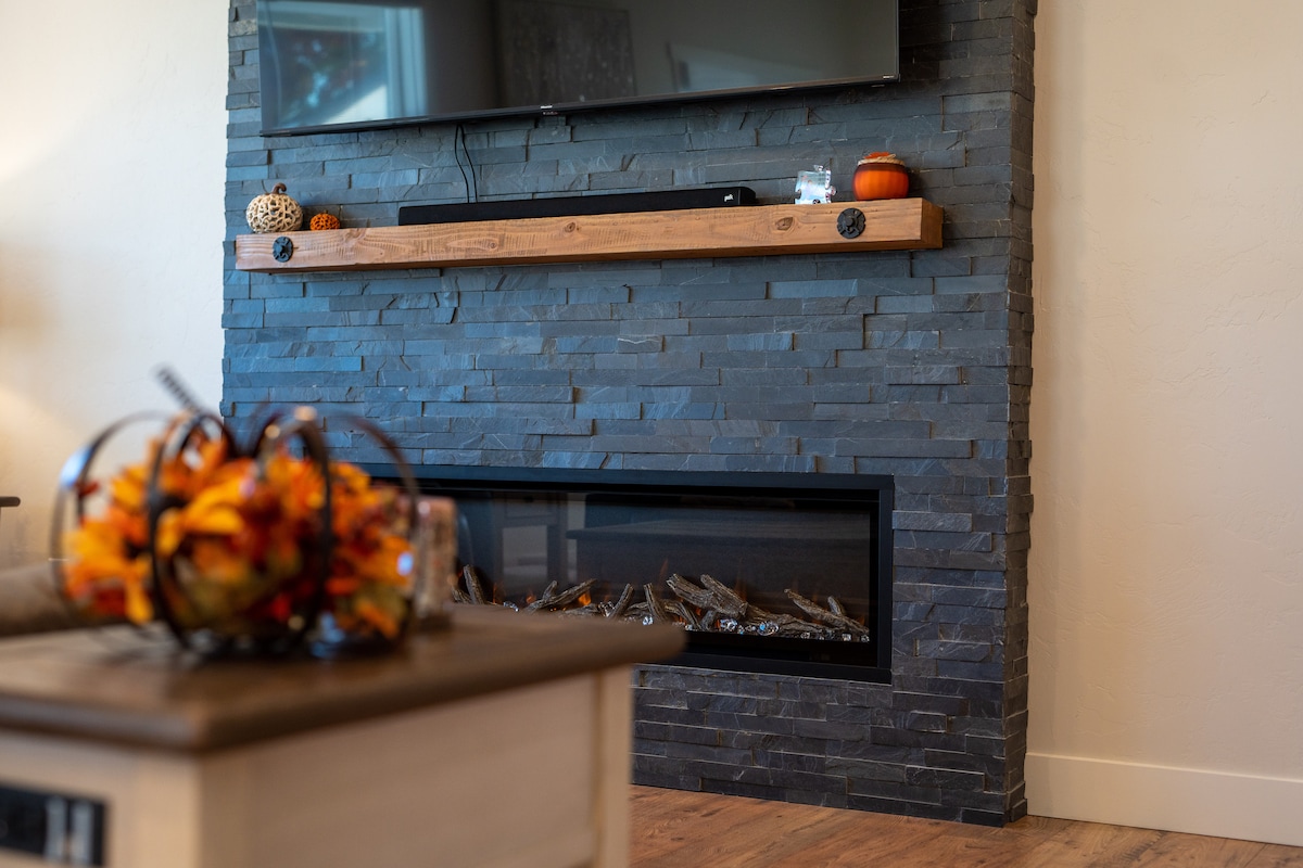 A modern living area features a sleek stone fireplace with a horizontal wooden mantel. Seasonal decorations, including small pumpkins, are displayed atop the mantel. A glass front reveals logs within the fireplace, while a decorative bowl of autumn leaves rests on a nearby table.