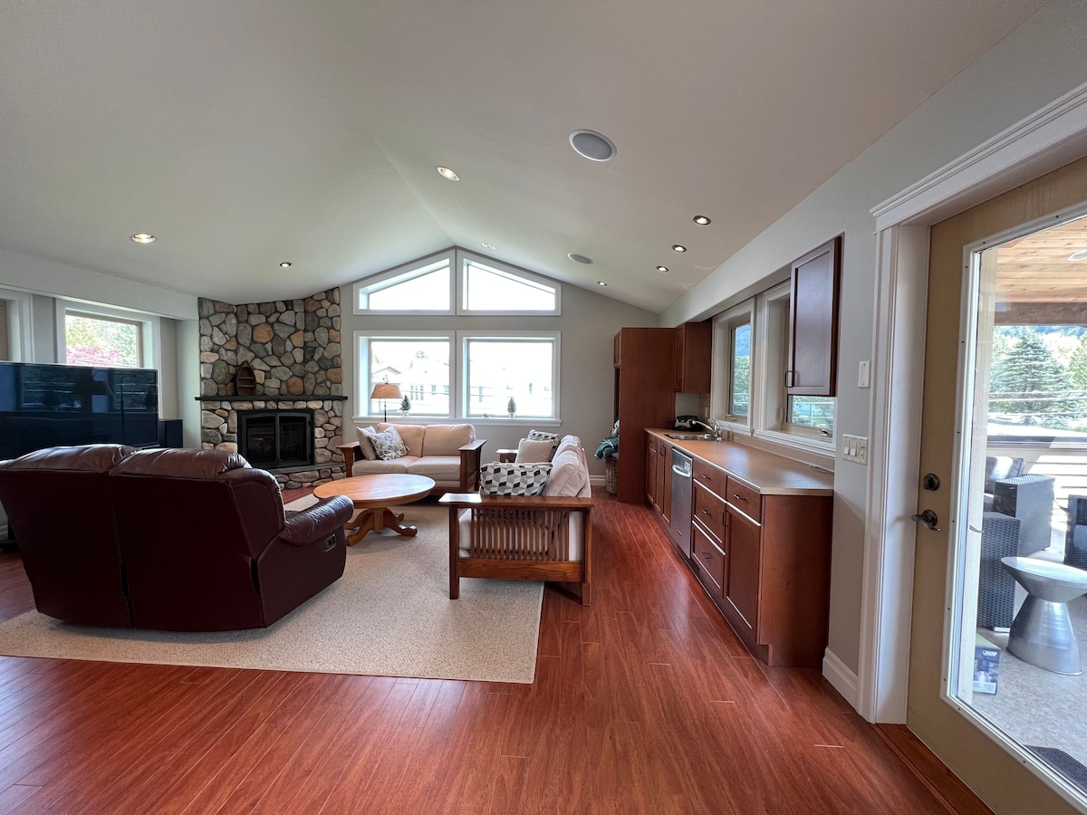 The open living area features a stone fireplace and large windows that allow natural light to fill the space. Comfortable seating arrangements include a leather recliner and a coffee table. A kitchen area with modern cabinetry and a patio door to the outdoor space are visible.