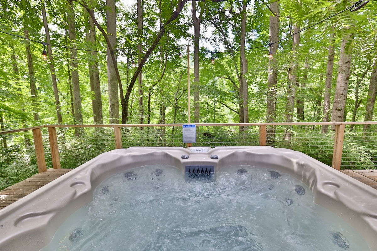 A spacious hot tub is situated on a wooden deck, surrounded by lush greenery. Tall trees create a natural backdrop, enhancing the serene atmosphere. The bubbling water in the hot tub invites relaxation, making it a perfect spot to enjoy the peaceful outdoors.
