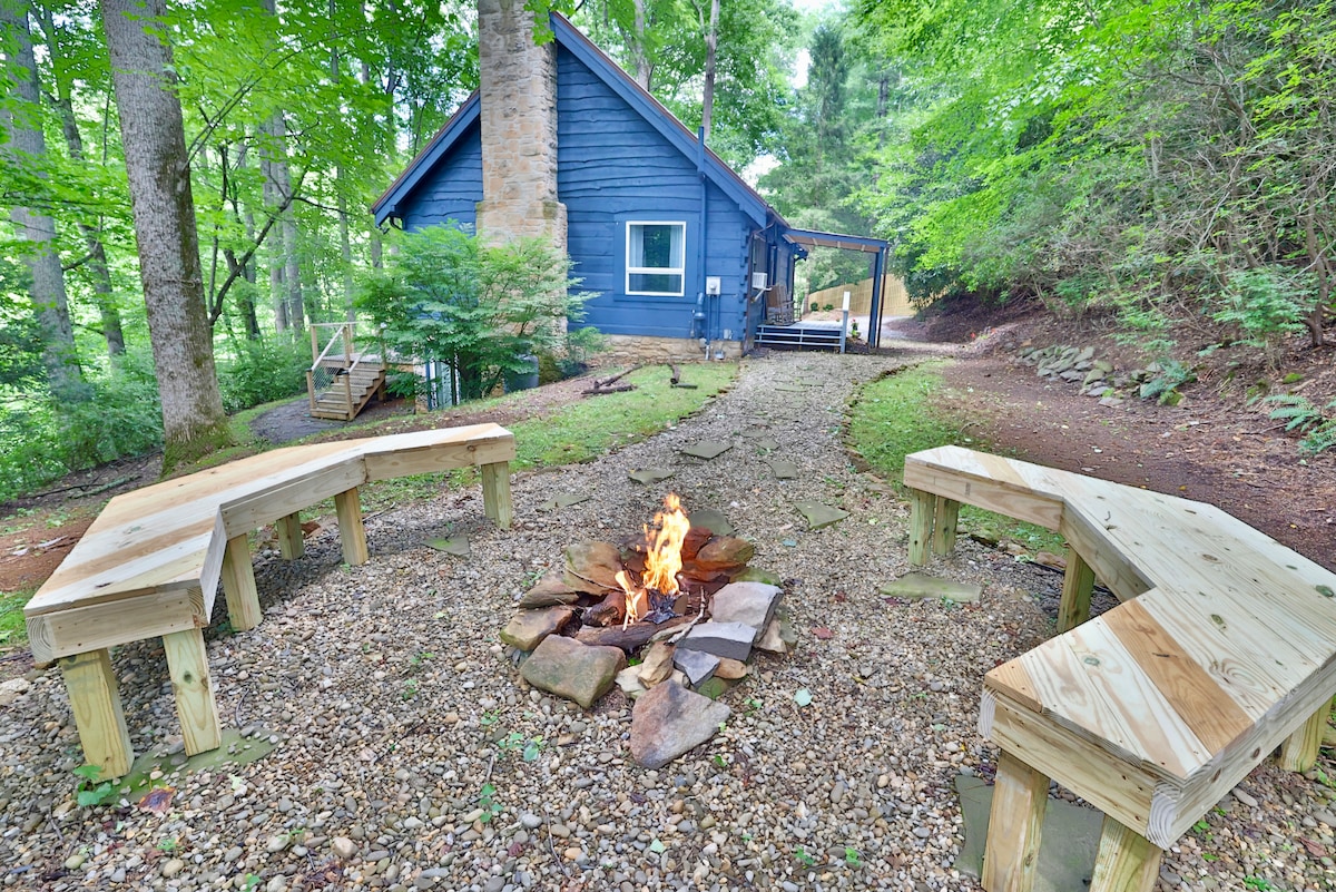 A fire pit area features circular benches constructed from wood, arranged around a gathering space of stones. The cabin, painted in deep blue, is partially visible in the background among trees and greenery, providing a serene outdoor setting.