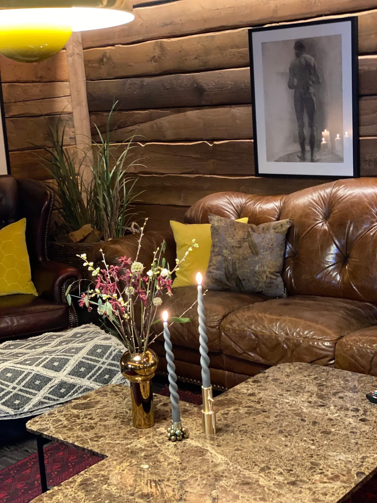 A cozy living area features a brown leather sofa adorned with patterned cushions. A decorative vase with flowers sits on a marble coffee table, accompanied by two lit candles. Neutral wooden walls offer a warm backdrop, enhanced by framed artwork.