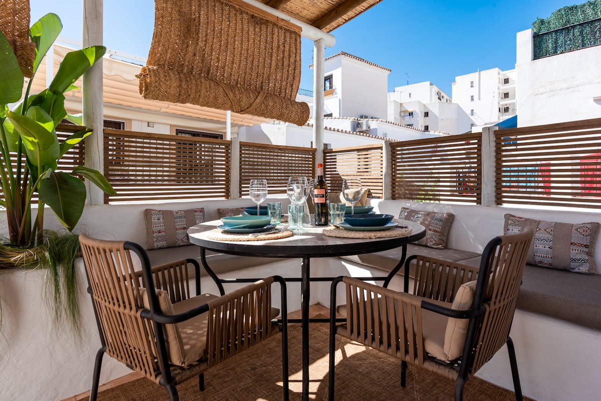 A dining area is nestled under a shaded canopy, featuring a circular table set with plates and glasses. Comfortable seating surrounds the table, complemented by decorative cushions. Lush greenery adds a touch of nature, while the backdrop reveals the vibrant architecture of the surrounding area.