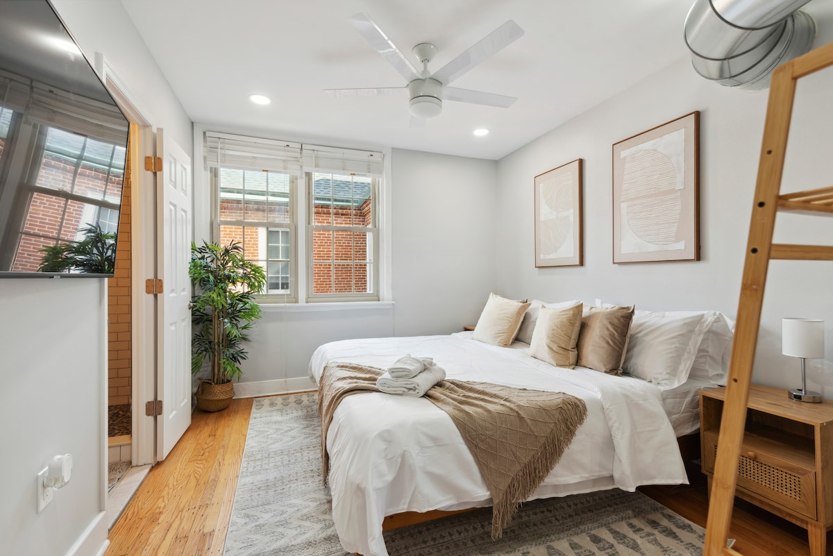 A serene bedroom is presented with a spacious bed adorned with neutral-toned pillows and a cozy blanket. Natural light filters through dual windows, highlighting the light-colored walls. A complementary ladder shelf and minimalist decor add to the room's inviting ambiance.